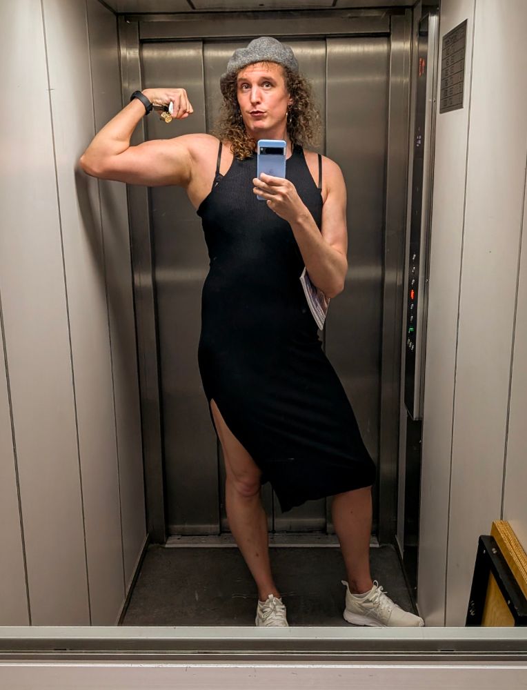 Alyx flexing her biceps in the mirror of the lift of her flat building. She's wearing a black dress with a leg split, white sneakers and a grey beret