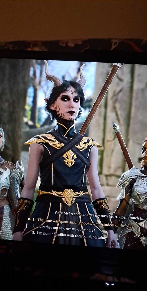 A screenshot of my player character in Baldurs Gate 3, a pale skinned Tiefling with black tendril neck tattoos, a black and gold outfit, black hair and glowing green eyes. She's basically the dream goth girl.  