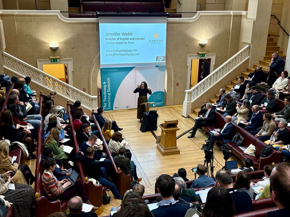 A lecture hall with leather rising seats. It is full of teachers - Jennifer Webb is giving a talk from the centre - her slides are projected on a large screen behind her.