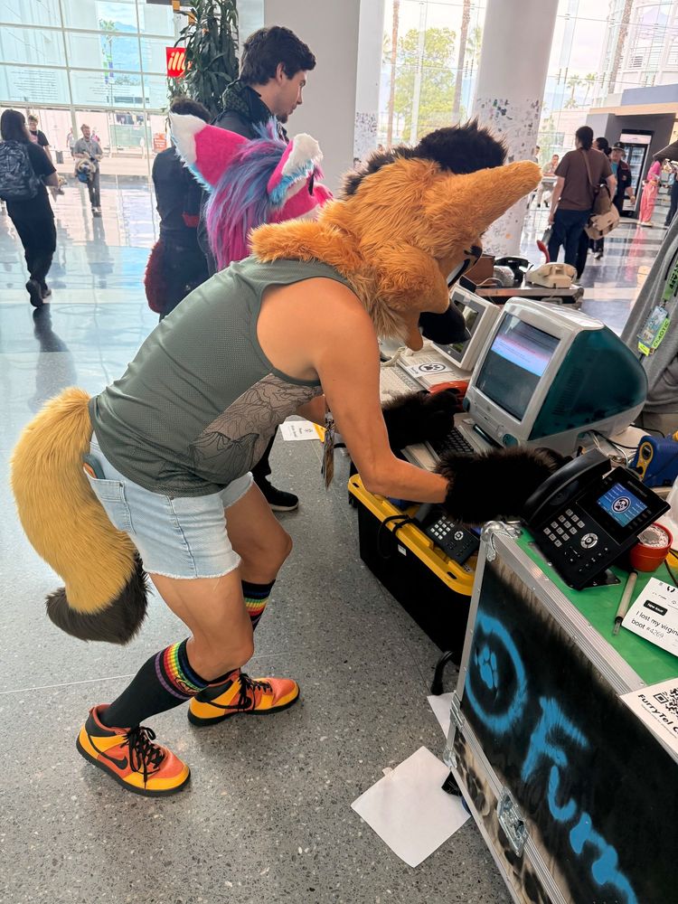 A person in a maned wolf fursuit head, yellow tail, rainbow knee-high socks, and orange sneakers is leaning over an old teal Apple iMac computer at a convention booth. They appear to be typing on the keyboard, with black paw gloves on their hands. The booth has various telephones, papers, and a large case with a paw print. Other attendees, including another person in a bright pink and blue fursuit head, can be seen in the background walking through the large, bright convention hall with glass walls.