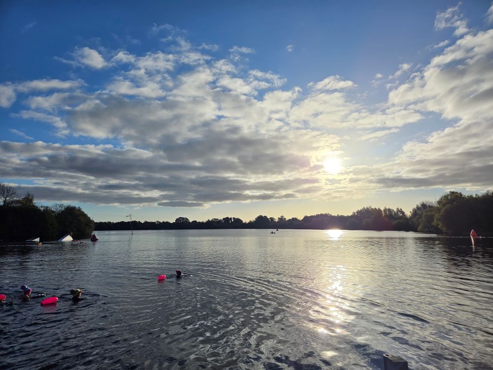 Sun poking out behind fluffy white clouds over a lake with people swimming