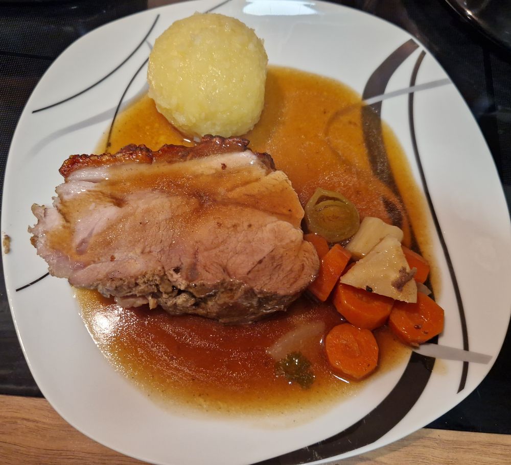 Ein angerichteter Teller mit einem Stück Schweinebraten, einer knusprigen Kruste, und brauner Bratensoße. Daneben liegt ein Kloß (Kartoffelknödel) und geschmortes Gemüse, darunter Karotten, Lauch und Sellerie.