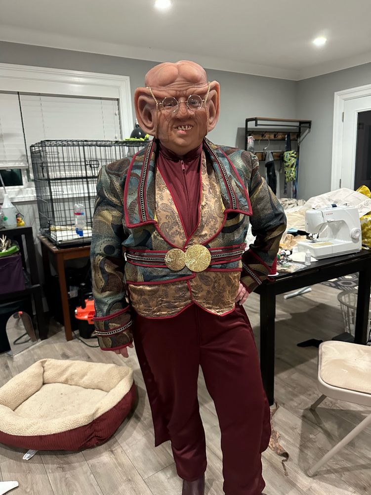 Me in my full Ferengi outfit so far. The base layer is a red speed suit. Over that is a gold paisley vest, and over that is a tailcoat made of a garish upholstery fabric. On my head, I’m wearing the prosthetic. I also have on glasses and pointed teeth 