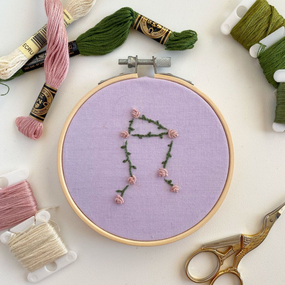 An embroidery of roses making the shape of the Libra constellation on a lavender background.
