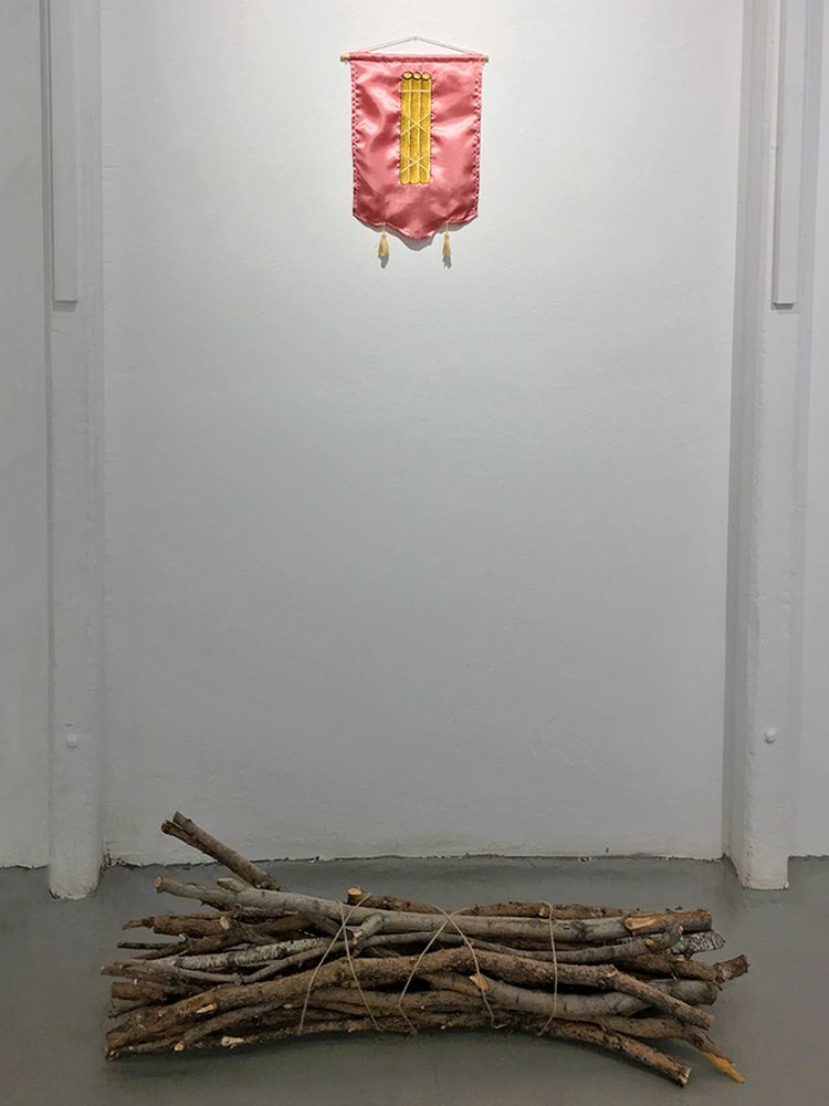 An embroidery of a bundle of sticks on pink satin fabric hanging above an actual bundle of sticks on the ground.