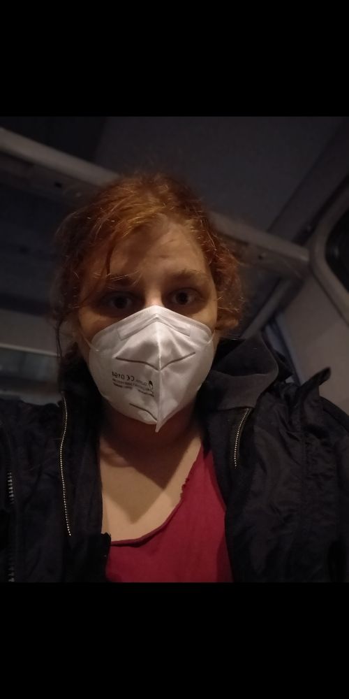 Ein Foto von mir mit Maske im Zug, wie ich in die Kamera schaue. Ich bin weiß, hab hennarote Haare, die zu einem unordentlichen Pferdeschwanz gebunden sind, buschige Augenbrauen und trage ein rotes Shirt mit ner schwarzen Jacke darüber.