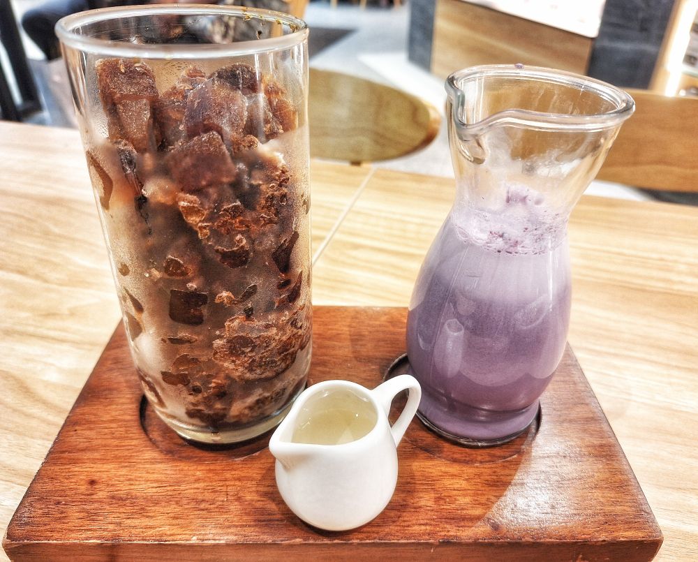 A glass full of cubed iced coffee and another glass with ube milk. A small container in front is filled with sweet syrup. 