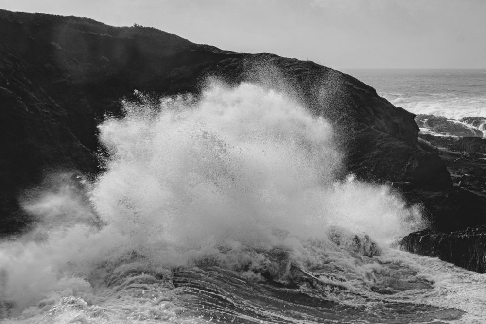 In black and white: crashing waves explode against a backdrop of jagged black rocks with the ocean horizon in the distance
