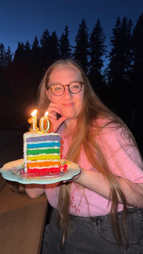 A photo taken outside after dusk. Bailey is holding a rainbow layered slice of cake with candles of the number 10 lit on top. She looks very happy and very proud. 