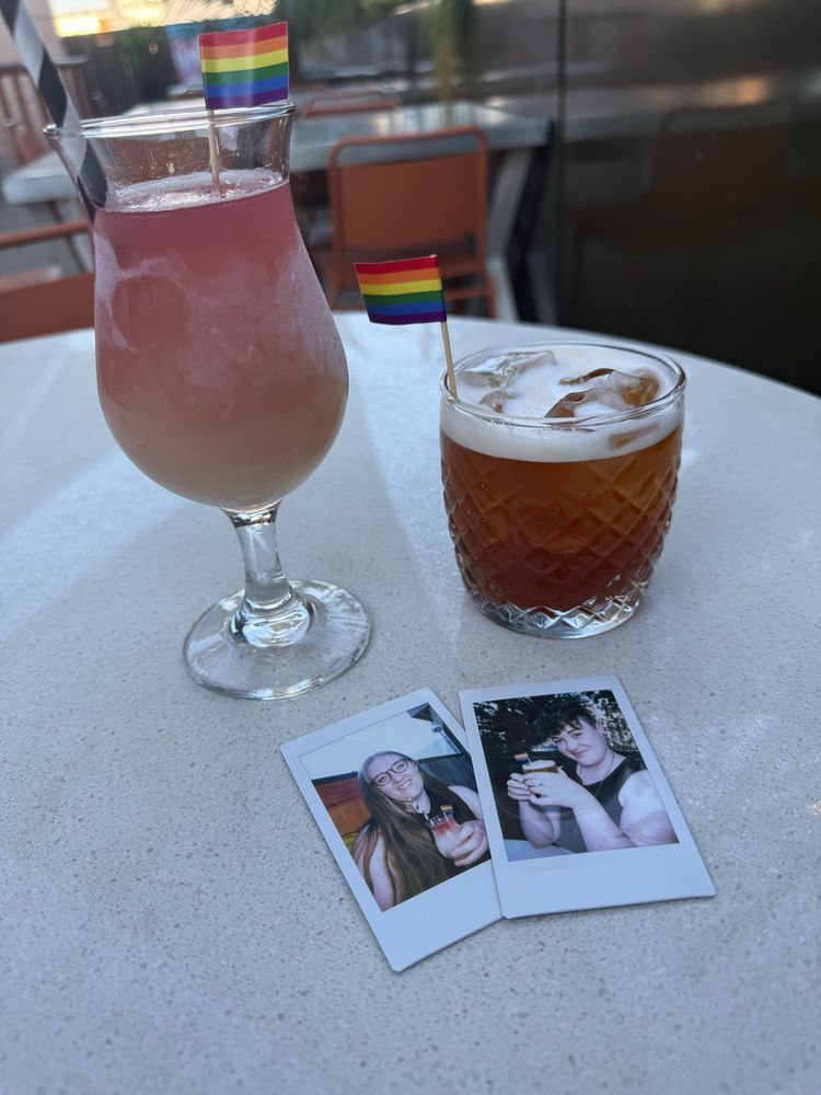 A shot of a table on a rooftop bar. There’s two drinks in the table with teeny tiny pride flag picks sticking out of them. In front of the drinks there are two Polaroid photos of Bailey and Alex holding those same drinks, they look (and are) very happy!
