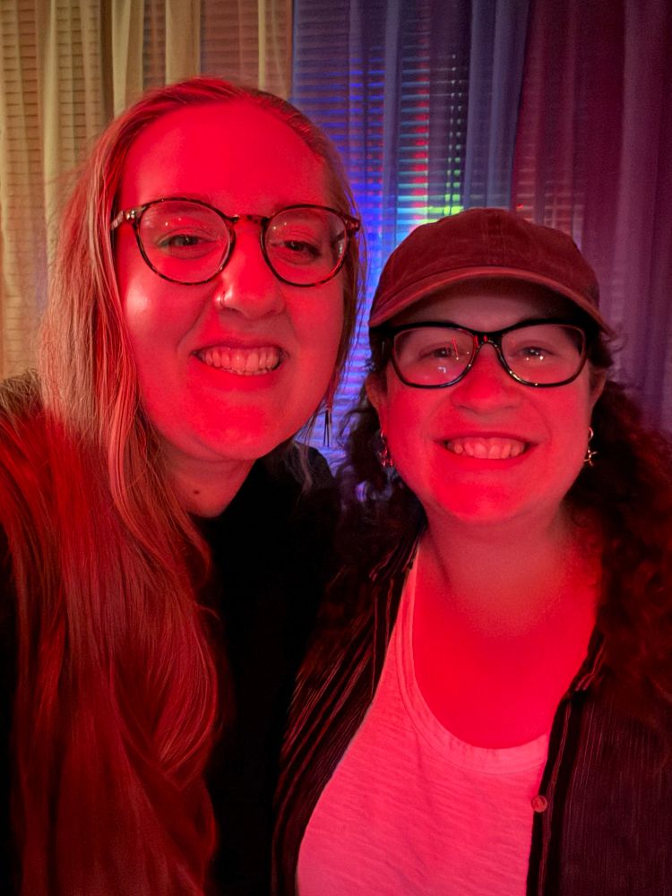 Bailey and Ashley smiling in red lighting, a rainbow curtain behind them 