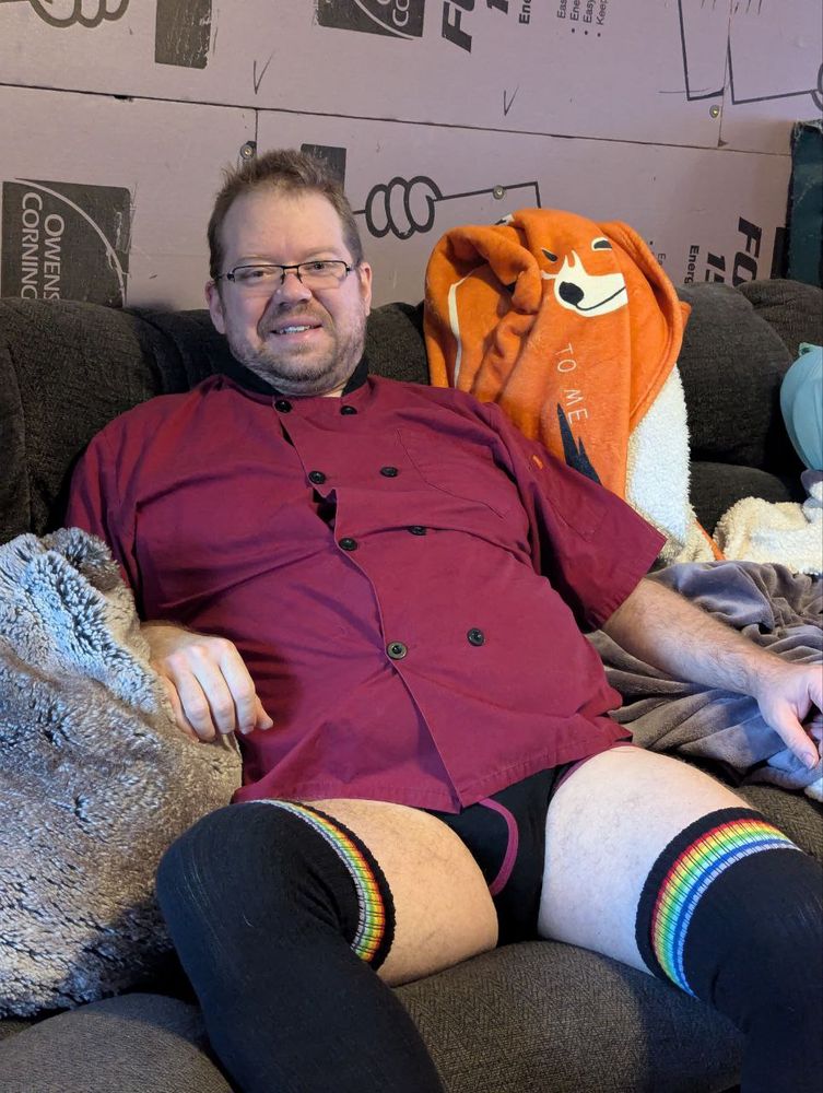 Freefox is sitting on a couch, getting ready for work in a burgundy chef coat with rainbow lined thigh-high stockings and black briefs with piping that matches his coat.