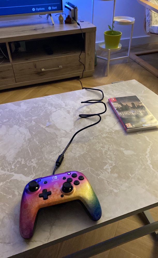 Multicoloured wired Nintendo switch controller together with Hogwarts legacy game on a table. In the background the Nintendo switch console.