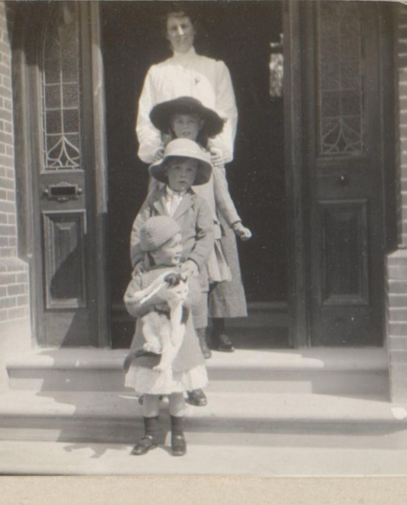 In this black-and-white photo, three girls and one woman stand at a doorway and on the stairs in front of the door. The smallest girl, in the front, holds a cat.