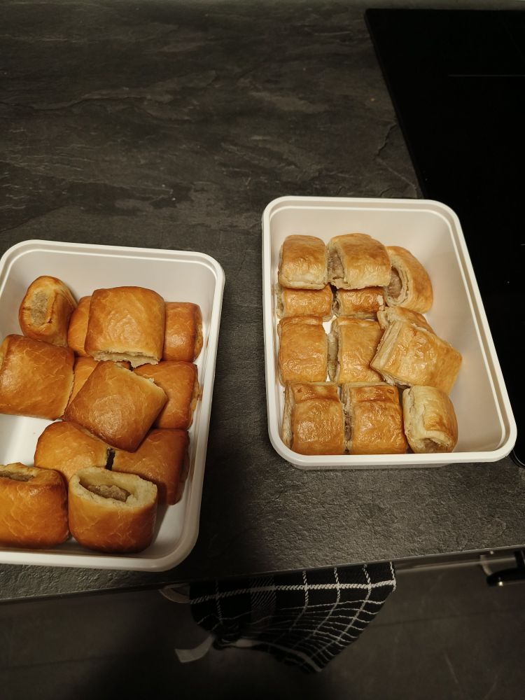 Picture of two plastic tubs with two different types of freshly baked sausage rolls on a Black-ish counter top.