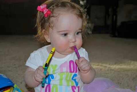 A toddler holding a yellow and a purple crayon, one in each hand, eating the purple one.