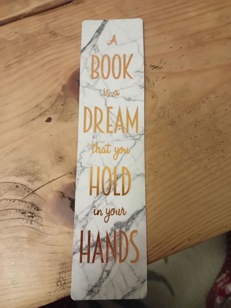 A bookmark on a wooden table that reads "A Book Is A Dream That You Hold In Your Hands". This quote is attributed to Neil Gaiman. This user does not support that vile little homonculus, and is choosing to believe the victims, so is throwing it out.