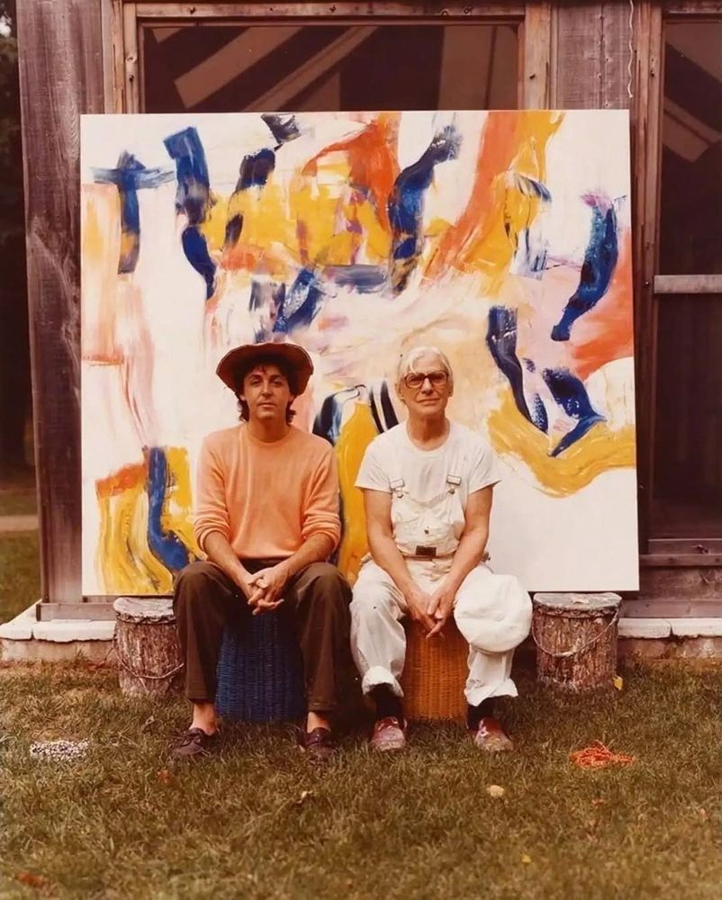 Paul McCartney and painter Willem De Kooning sitting in front of one of the latter’s paintings while outdoors.