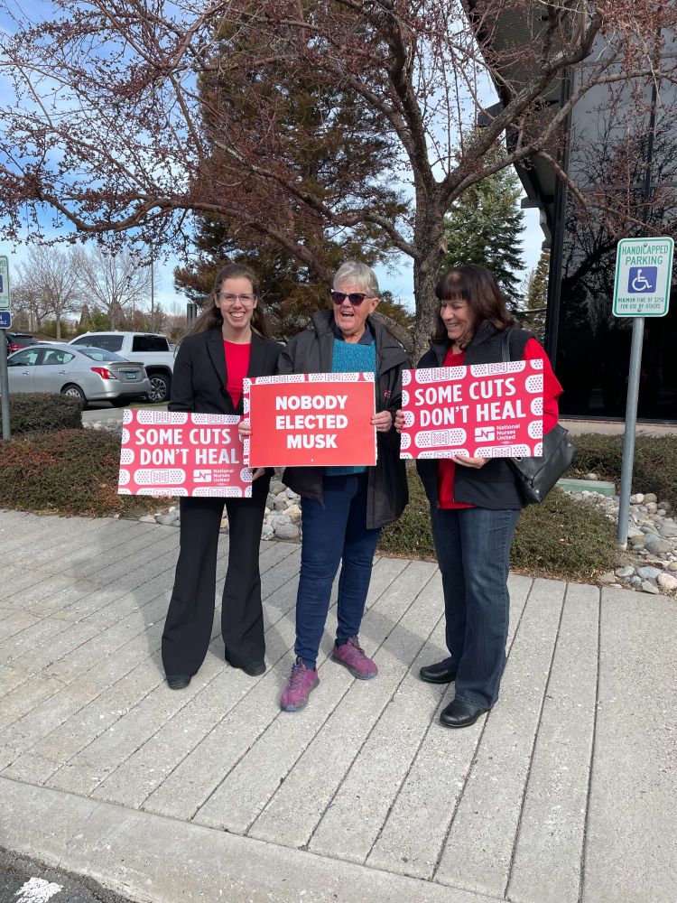 Three people holding protest signs outside a building. The signs advocate against budget cuts, specifically mentioning "Don't Cut Medicaid."
