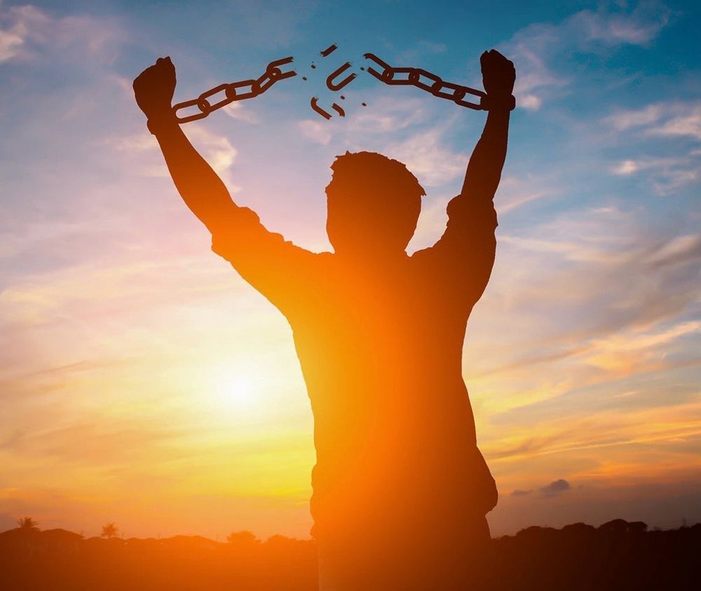 silhouette of a man raising his arms above his head and breaking a chain binding his hands in front of a sunset