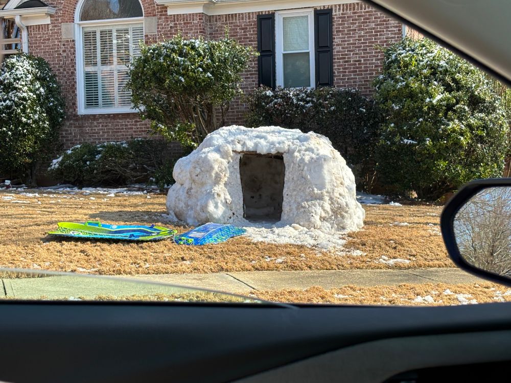 A snow igloo made in someone’s front yard in Georgia after the snowstorm on January 10; two sleds lie on the clear grass next to it with the bushes having light snow on them.