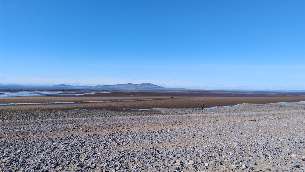 Looking across the Solway Firth towards Criffel in Scotland. It's low tide and the sea is barely visible beyond a long stretch of sand and mudflats, with only two small figures in the distance. The sky is brilliant blue.