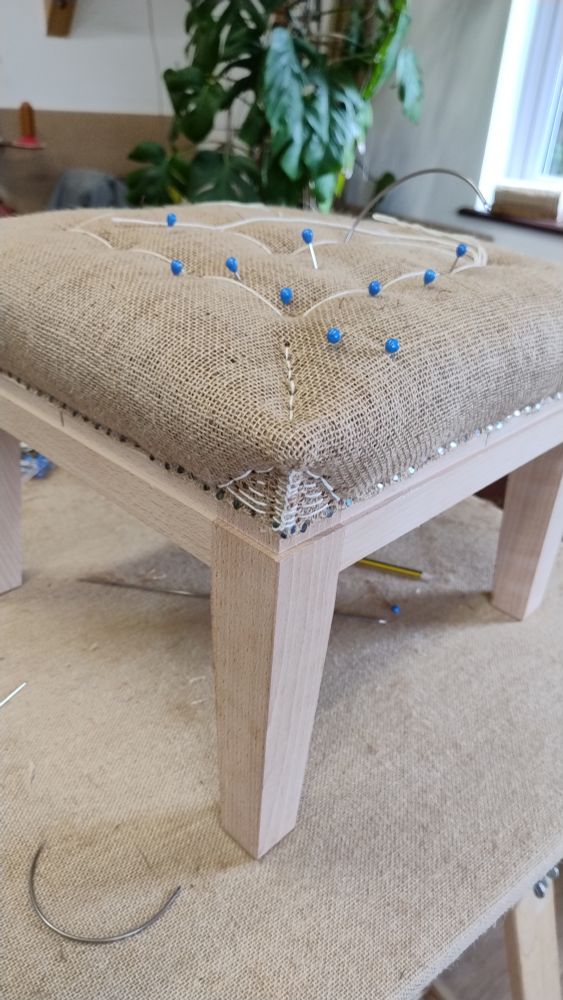A foot stool with linen scrim covering the stuffing. The scrim has been stitched in the corners at the legs to give them a sharp edge. The top of the stool has been marked with blue pins to guide the stitching 
