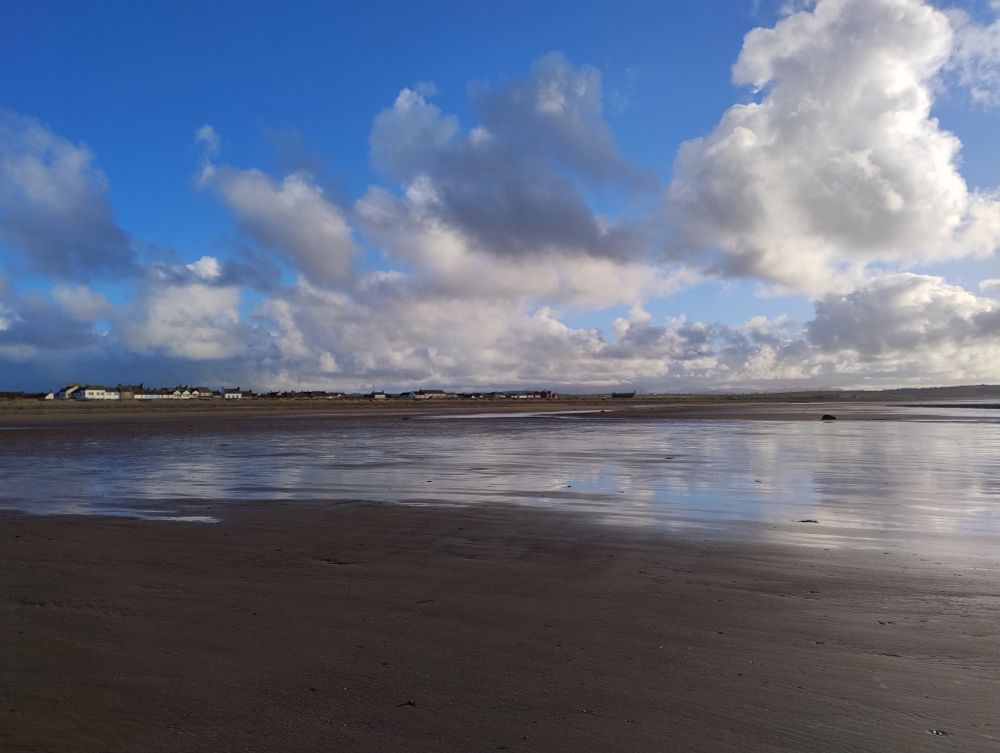 Blue sky and white clouds reflected in a pool of water on a large flat sandy beach at low tide.