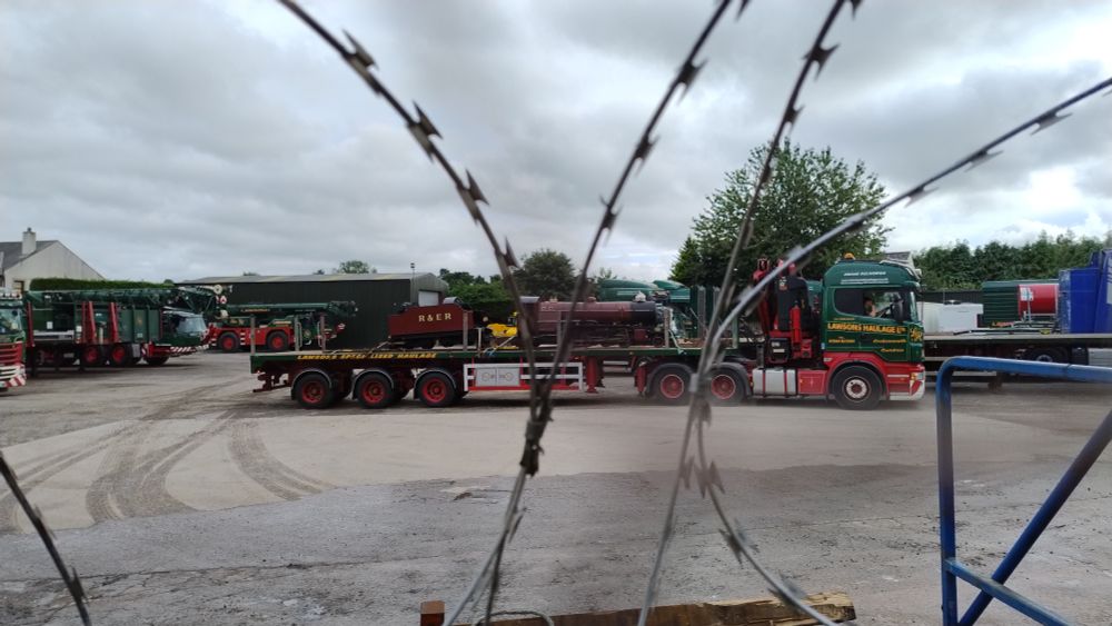 Looking through razorwire into a haulage yard. On a low loader lorry, decorated in the green red and gold of Lawson's Haulage livery, there is a small, narrow guage steam engine and coal tender. The engine and tender are painted burgundy, and have the initials R & ER painted on them for Ravenglass and Eskdale Railway.