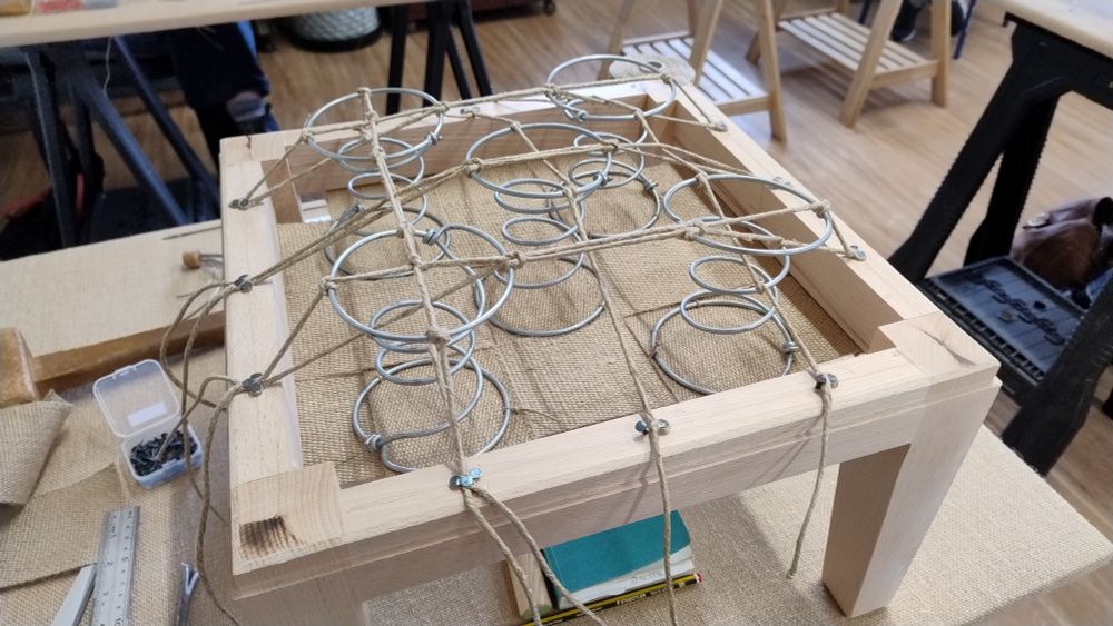 A square footstool in the process of being upholstered , with five coil springs tied to the wooded frame with strong cord