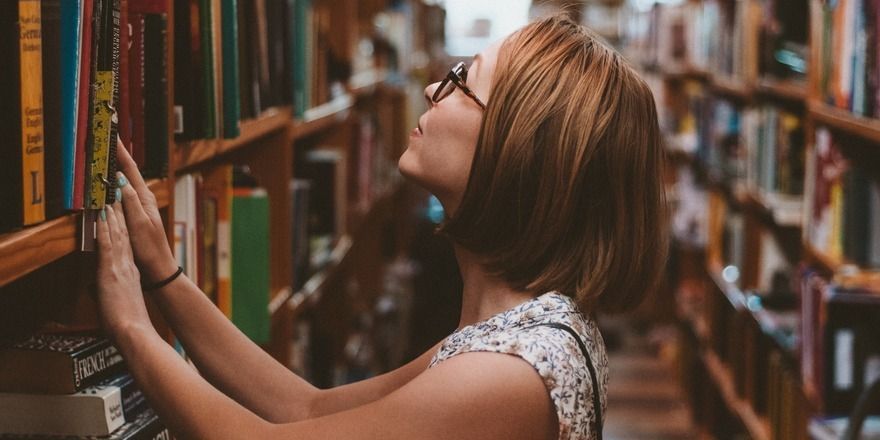 A brunette woman with glasses rests her hands against bookshelves as she gazes up at a higher shelf.