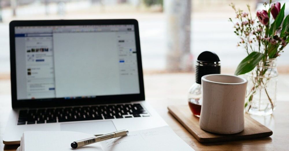 A laptop with several windows open on it is seen next to a mug and a small vase of flowers.