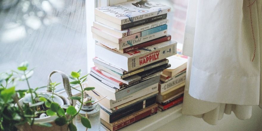 Paperbacks are stacked in a windowsill between a few small plants and white curtains.