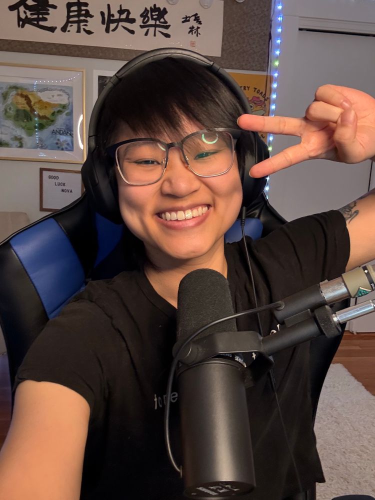Connie grins and holds up a peace sig in a selfie. They are a Chinese person with short black hair, over the ear headphones, and a black T-shirt. They sit in a blue and black gamer chair behind a black Shure SM7B microphone.
