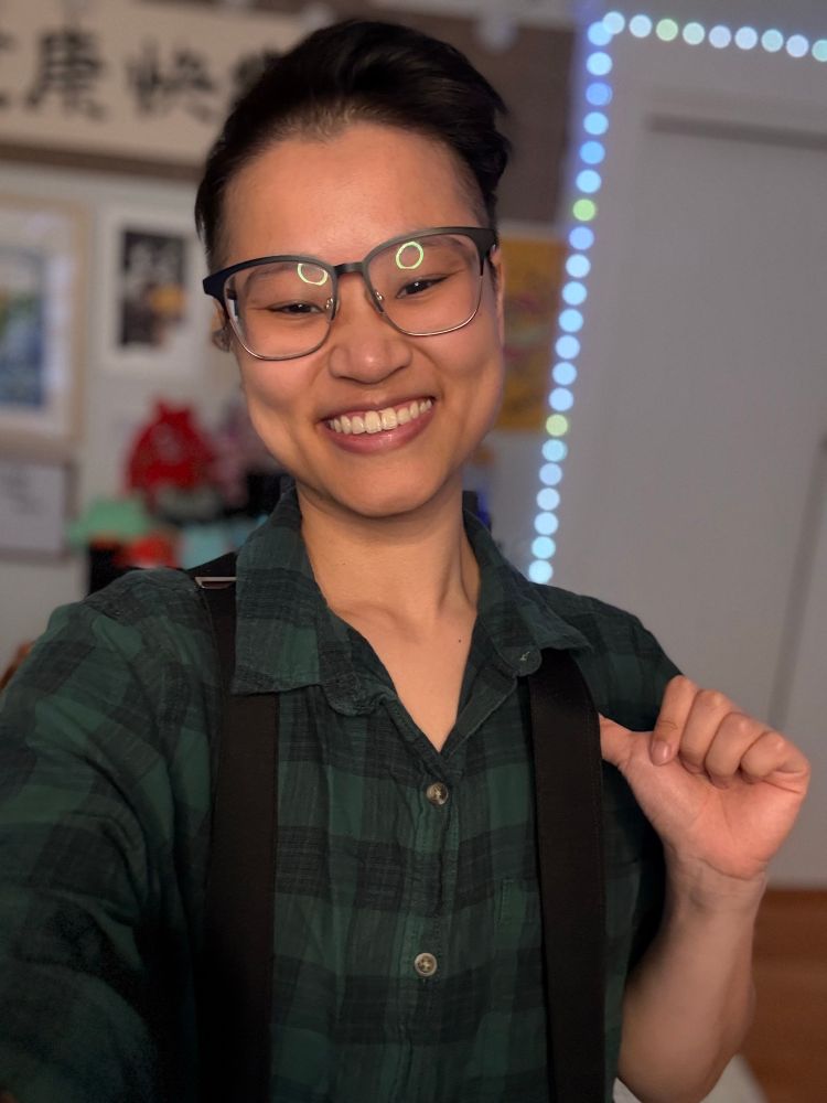 Connie grins against a blurry background of their home office. They are a Chinese person with short black hair shaved on the sides and styled up. They wear square black glasses, black suspenders, and a dark green flannel shirt. Their left thumb is hooked under a suspender as they pose cheekily for the camera.