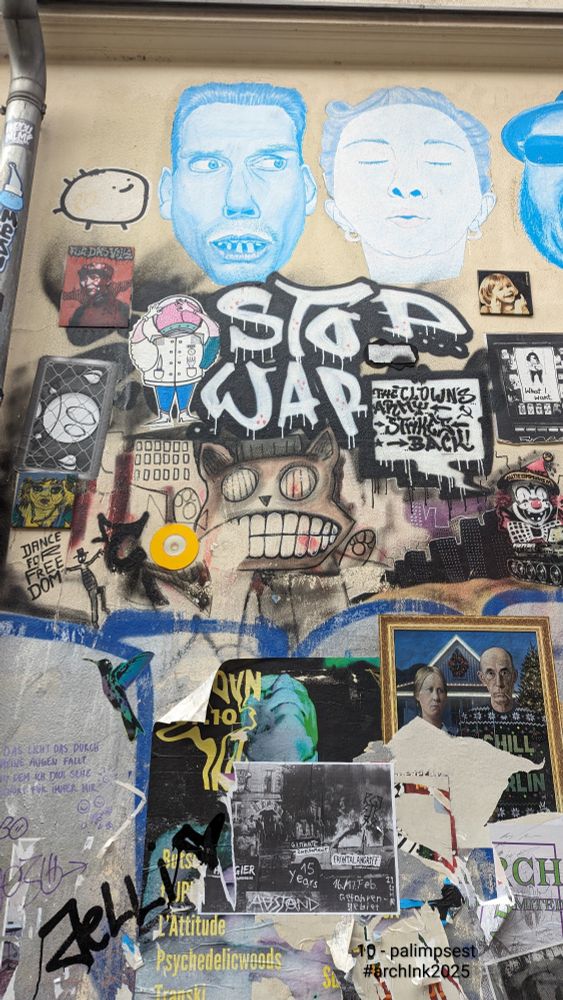 A city wall layered with street art, stickers, and posters. Blue-painted faces appear above, with bold text reading “STOP WAR” and various smaller artworks below. Older posters peek through torn edges and paint drips.
