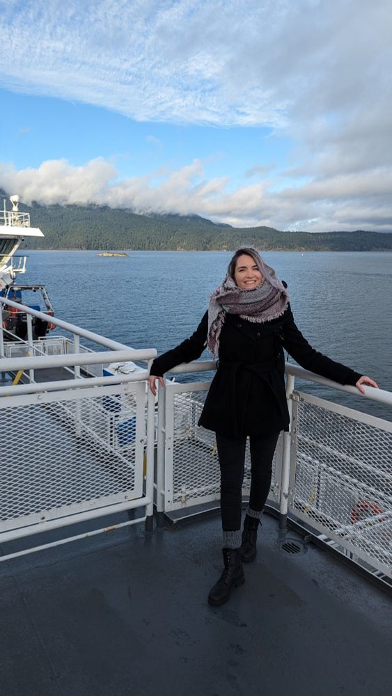 Jona, a woman stands smiling on the deck of a ferry on the way to Victoria BC, wrapped in a scarf and black coat. Behind her, calm waters stretch out toward forested mountains under a blue sky with wispy clouds. The atmosphere is crisp and bright, suggesting a winter or early spring day.