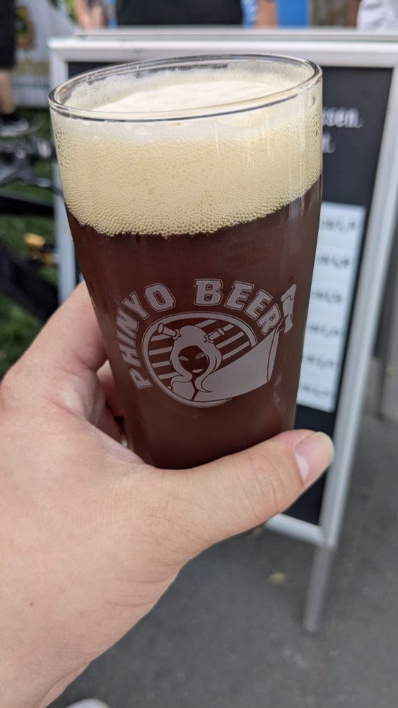 Ich halte ein Glas dunkles Bier mit dem Branding von "Phinyo Beer"