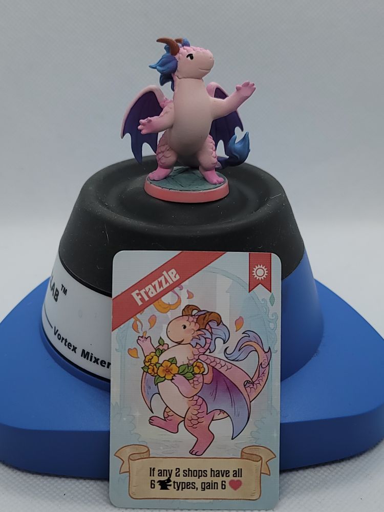 A painted miniature from the Flamecraft board game sitting on top of a Vortex Mixer. Propped up below it is the card from the board game that the paint scheme of the miniature is based on, the fancy dragon named Frazzle. It is a small pink dragon with a lighter pink stomach, with wings and tufts of fur that fade from purple to light blue.
