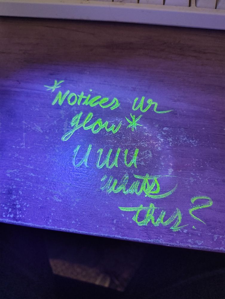 The same desk but under UV light, saying "*notices ur glow* uwu what's this?"