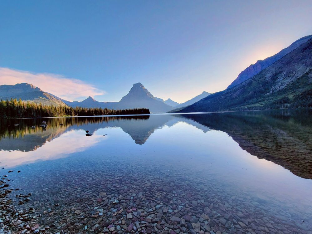 Stunningly beautiful and peaceful Two Medicine Lake in Glacier National Park. The mountains that embrace the lake and the cloudless sky are reflected in the deep stillness of the alpine body of water. The multicolored stones that make up the lake bed can be seen in the shallows at the bottom of the picture. 