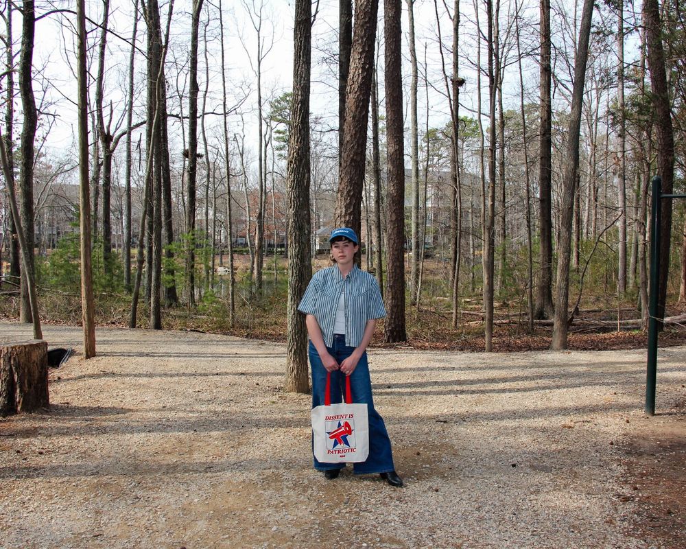 Image of a person in the woods wearing a blue hat that says "Dissent" and holding a tote bag that says "Dissent is patriotic."