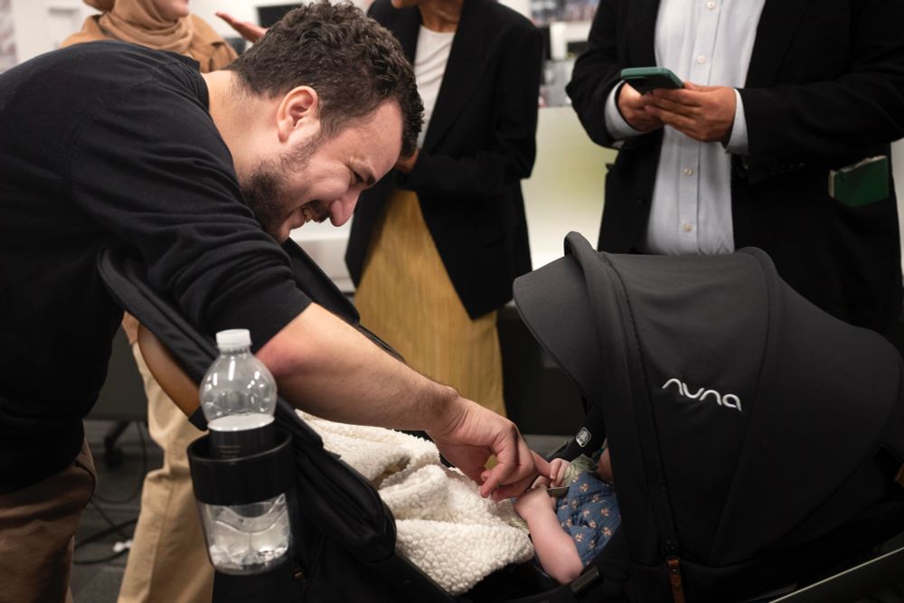 A photo of Mahmoud Khalil smiling and bending over a black stroller, with a baby’s small hand sticking out and grabbing Mahmoud’s finger. 