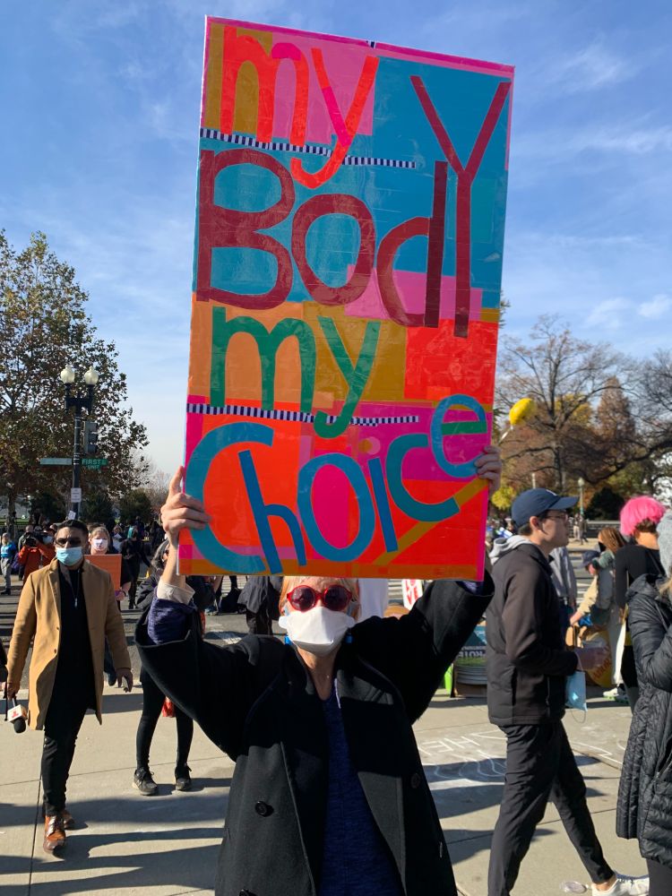 A person in a black coat stands outside holding with a colorful sign about their head reading "my body my choice" among a crowd.