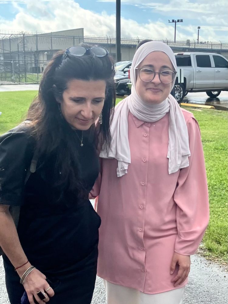 Two woman stand next to each other and hold onto one another. The woman on the left is wearing a black dress. The woman on the right is smiling and wearing a white hijab, glasses, and a pink garment. In the background is the fence of an ICE detention center.