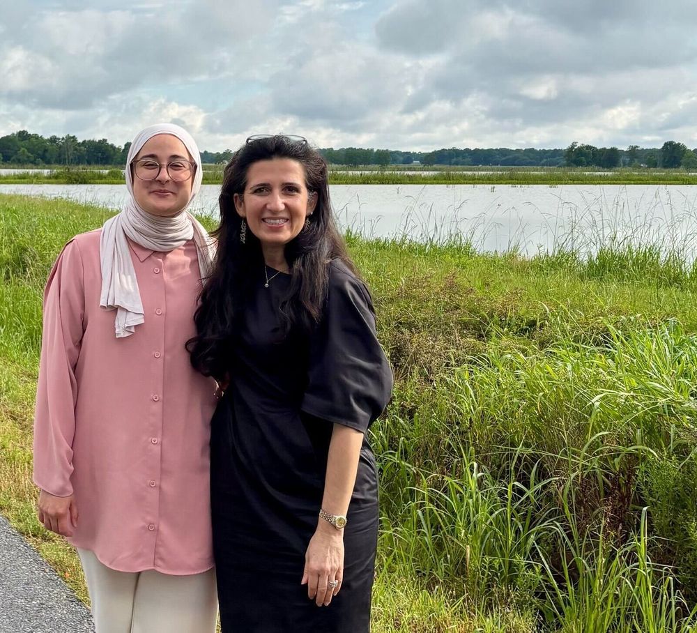 Two women standing, smiling, and embracing each other with grass, water, and the sky in the background. The woman on the left is wearing a white hijab, glasses, and a pink garment. The woman on the right is wearing a black dress.