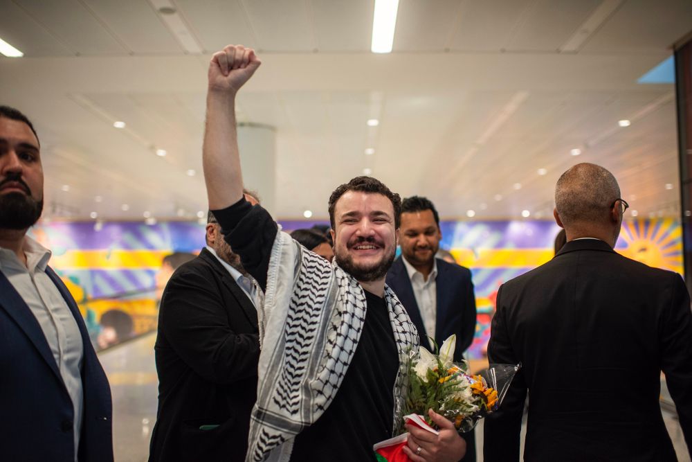 A photo of Mahmoud Khalil holding up a fist and smiling wide, and holding a bouquet of flowers. He is wearing a Keffiyeh. 