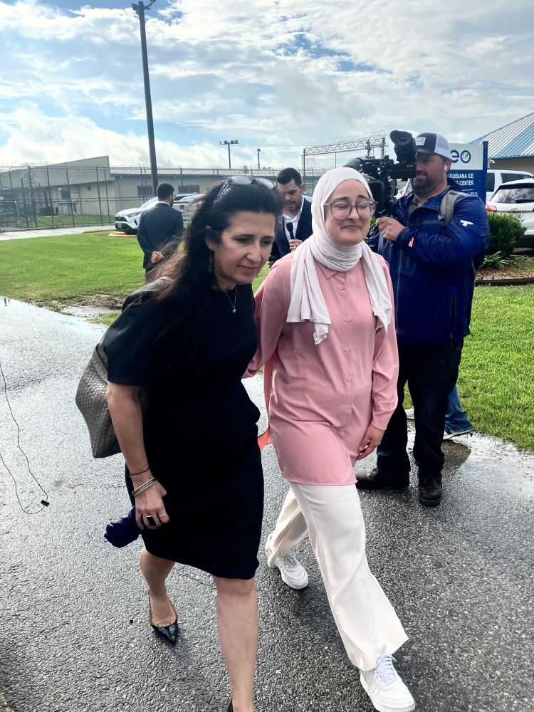 Two woman walking and embracing each other in front of an ICE detention center. The woman on the left is wearing a black dress and heels. The woman on the right is smiling and wearing a white hijab, glasses, and a pink garment. Members of the press are in the background.