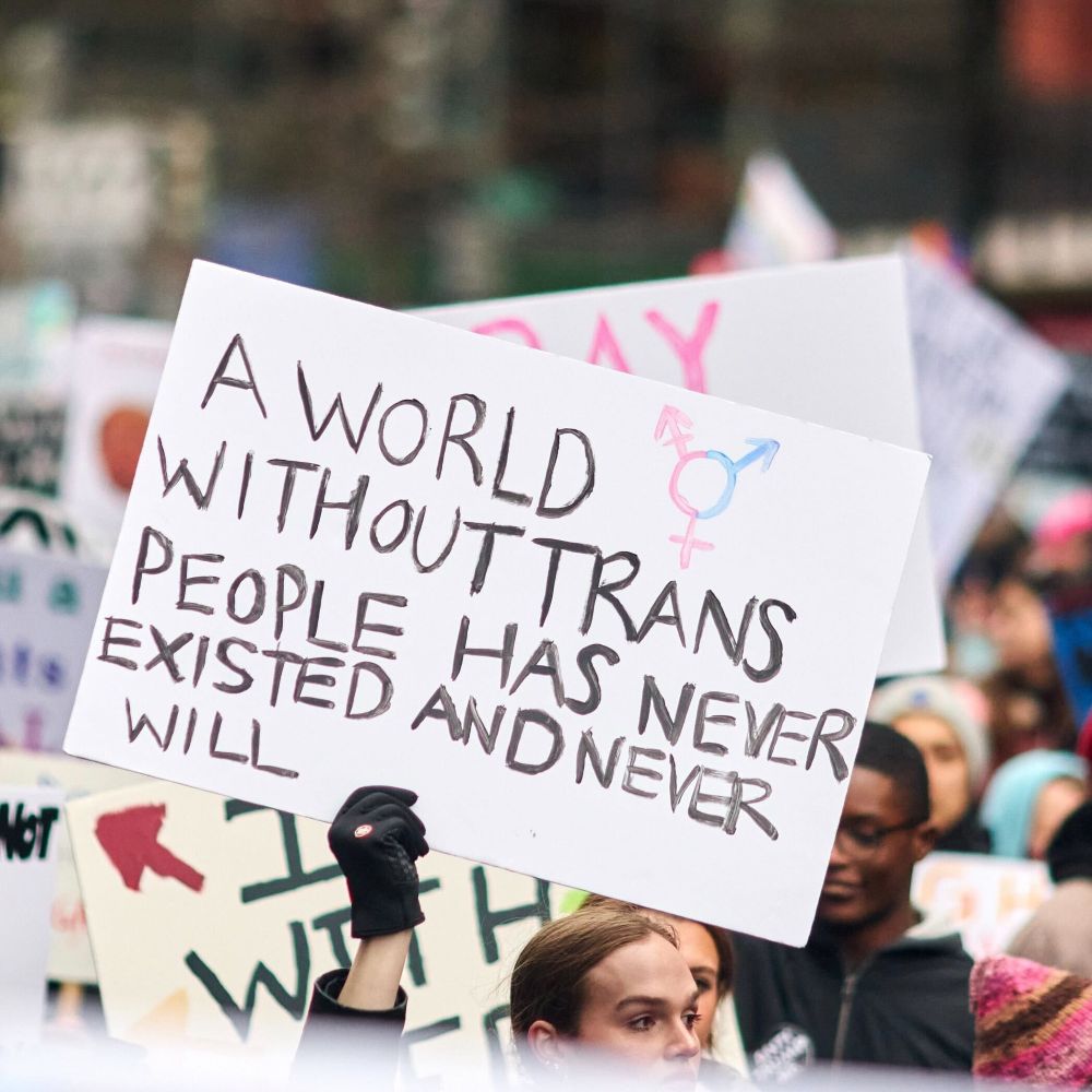 A photo of a person holding up a handmade rally sign reading "A world without trans people has never existed and never will" among a group of protesters.
