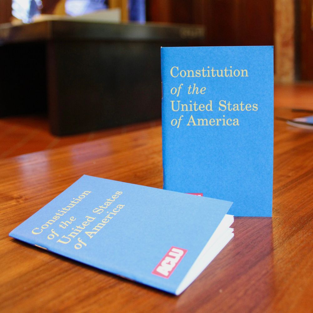 Photo of two blue pocket-sized Constitutions that read "Constitution of the United States of America" in yellow text on each of them with a red ACLU logo, on a wooden surface.